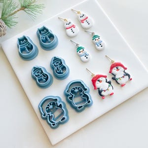 May include: A set of 10 blue and white polymer clay earrings featuring snowman and penguin designs. The earrings are displayed on a white surface with blue cookie cutters.