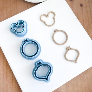 May include: A set of five cookie cutters, two blue and three brown. The blue cutters are shaped like a Mickey Mouse head, a Christmas ornament, and a Christmas tree. The brown cutters are shaped like a Mickey Mouse head, a circle, and a teardrop.