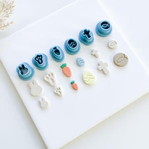 May include: A set of blue plastic cookie cutters and fondant decorations for Easter. The cutters include shapes of a bunny, a cross, a fish, a paw print, an egg, and a carrot. The fondant decorations include a bunny, a paw print, a carrot, an egg, a cross, and a fish.