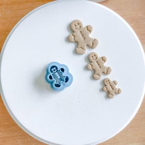 May include: Three gingerbread man cookie cutters, one blue and two brown, on a white surface.