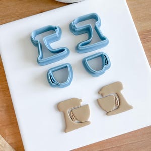 May include: A set of blue and brown plastic cookie cutters in the shape of a stand mixer and a cup. The cutters are on a white surface.