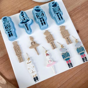 May include: A set of nine nutcracker and ballerina themed earrings. The earrings are made of polymer clay and feature a variety of colors, including brown, white, pink, blue, and green. The earrings are all different shapes and sizes, and some have glitter accents.