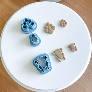 May include: A set of six blue plastic cookie cutters in the shapes of a turkey, a paw print, a chicken, and a bird. There are also two brown clay cutouts of a chicken and a paw print.