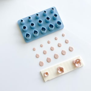 May include: A blue silicone mold with 16 different sized teardrop shapes. There are 16 pink teardrop shapes and 3 pink flowers on a white surface.