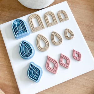 May include: A set of 10 polymer clay cutters in various shapes and sizes. The cutters are arranged on a white surface. The cutters are in shades of blue, brown, and pink.