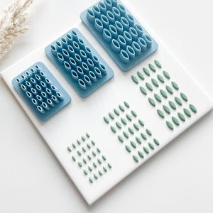 May include: A top-down shot shows three blue oval-shaped cutters in different sizes, alongside green oval-shaped clay pieces. The cutters are arranged on a white surface, with the clay pieces next to them. The cutters are for crafting.