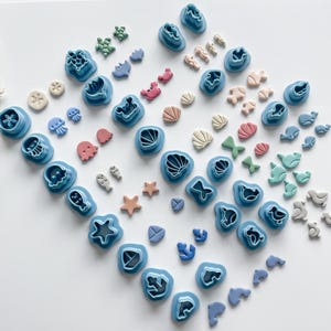 May include: A set of blue plastic cookie cutters in various ocean-themed shapes, including seashells, starfish, fish, jellyfish, and a crab. The cutters are arranged on a white surface with small, colorful clay shapes.