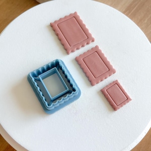 May include: Three different sized cookie cutters, two are pink and one is blue. The blue cutter is a double-layered frame design. The pink cutters are simple rectangular shapes with a scalloped edge.
