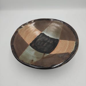 May include: A shallow, round ceramic bowl featuring a segmented design with a central black square surrounded by alternating panels of muted brown, tan, and sage green. The surface has a glossy, glazed finish with visible texture and variations.