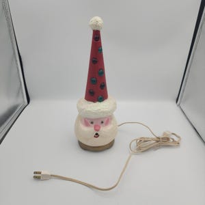May include: A vintage ceramic Santa Claus lamp featuring a tall, conical red hat adorned with embedded green and dark-colored circular lights. The figure has a textured white beard, a pink face, and a beige base, connected to a white electrical cord.