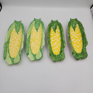 May include: Four ceramic corn-shaped serving dishes. Each dish features a yellow corn cob design with green husks. The dishes are arranged on a white surface, showcasing their detailed, hand-painted appearance.