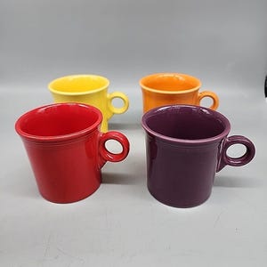 Könnte beinhalten: Ein Set aus vier Keramikbechern in den Unifarben Rot, Gelb, Orange und Lila. Jeder Becher hat eine glatte, glänzende Oberfläche, einen breiten kreisförmigen Rand und einen kleinen, runden seitlichen Henkel. Die Becher sind in einer Zweierreihe angeordnet.