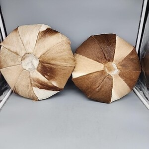 May include: Two round decorative pillows. Each pillow is made of sections of brown, tan, and cream-colored animal hide. The pillows have a central, circular design and a textured, natural appearance. The pillows are placed on a gray surface.