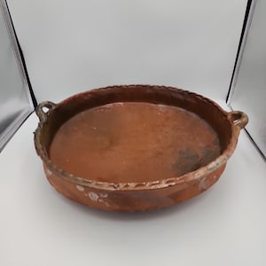May include: A round, shallow terracotta baking dish with two small side handles. The exterior features a reddish-brown finish with subtle variations in color and a textured, scalloped rim detail along the top edge of the vessel.