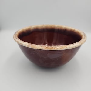 May include: A round ceramic bowl with a glossy dark brown interior and exterior finish. The rim features a thick, textured band of cream-colored glaze with brown speckling that creates a distinct contrast against the solid dark brown body.