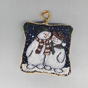 May include: A square tapestry Christmas ornament featuring two snowmen with red and brown scarves and hats, set against a dark blue snowy background. The ornament has a gold rope loop for hanging and a gold border.