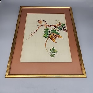 May include: Framed artwork featuring a bird perched on a branch with green leaves and yellow flowers. The artwork is set against a light background with a pink mat, enclosed in a gold-colored frame. The bird has brown and white feathers.
