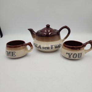 May include: A ceramic tea set featuring a teapot and two matching cups with a brown and cream gradient glaze. The teapot is inscribed with the text TEA FOR TWO, while the two cups are individually labeled with the words ME and YOU in dark lettering.