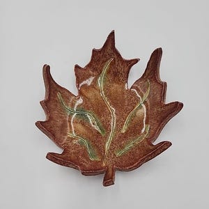 May include: A ceramic leaf-shaped dish in shades of brown, with green and white accents. The dish has a textured surface and is designed to resemble a maple leaf. The dish is likely used for decorative purposes or serving small items.