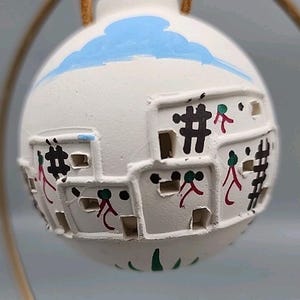 May include: A white ceramic ornament featuring a village scene with square buildings and cut-out windows. The ornament has a blue sky design at the top and is decorated with black, red, and green details. It hangs from a gold-colored loop.
