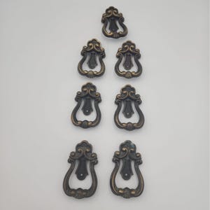 May include: A set of seven matching antique brass keyhole escutcheons arranged in a vertical pattern. Each escutcheon features a decorative scrolled top, a central teardrop-shaped opening for a key, and a curved, ornate metal frame finish.