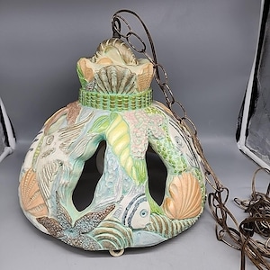 Może przedstawiać: Ceramiczna lampa wisząca o tematyce morskiej. Oprawa pomalowana jest pastelowymi kolorami i posiada rzeźbione muszle, rozgwiazdy i ryby. Wisi na łańcuchu w kolorze brązu.