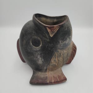 Puede incluir: Un recipiente de cerámica de color gris oscuro con forma de pez. La pieza presenta una abertura ancha en la parte superior, una hendidura circular para el ojo y detalles de aletas grabados con reflejos de color marrón rojizo en los bordes.