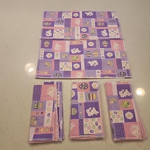 May include: An Easter-themed table setting with a large placemat and several folded napkins. The fabric features a patchwork design with bunnies, Easter eggs, and floral patterns in shades of purple, pink, and white.