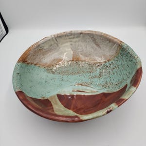 May include: A round ceramic bowl featuring a multi-toned glaze with a smooth, glossy finish. The interior displays a blend of earthy brown, creamy white, and a textured, seafoam green section with a crackled, organic pattern across the surface.