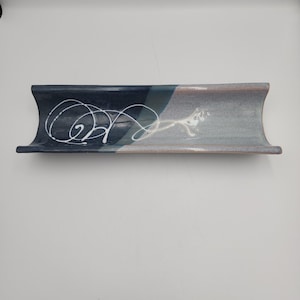 May include: A rectangular ceramic tray with a concave, curved profile. The surface features a two-tone glaze, transitioning from a deep navy blue on the left to a light, speckled grey on the right, accented by a white, swirling line design.