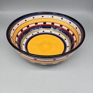 May include: A decorative ceramic bowl with a vibrant, striped design. The bowl features bands of yellow, red, blue, and green, accented with black squares and diamond shapes. The interior is a solid yellow, and the rim is black.