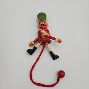 May include: A wooden nutcracker ornament featuring a green hat, a red jacket with gold accents, and a painted face with a black mustache. The figure has movable limbs attached to a red string that extends downward, ending in a small red wooden bead.