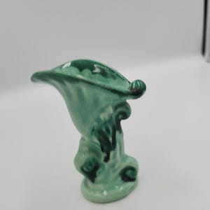 May include: A small, ceramic vase in a seafoam green colour. The vase has a flared top and a decorative, swirling base. The glaze has a glossy finish. The vase is a decorative item.