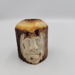 Puede incluir: Una escultura cilíndrica tallada con un rostro humano. La escultura está hecha de un material marrón claro con un exterior marrón más oscuro. La cara está tallada en un color más claro, con detalles. La parte superior de la escultura es de color marrón claro.