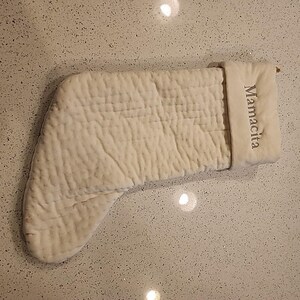 Puede incluir: Calcetín navideño color crema con textura acolchada. El puño del calcetín está bordado con la palabra "Mamacita" en fuente gris. El calcetín está hecho de un material suave y afelpado.