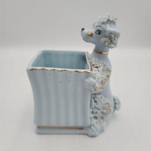 May include: A light blue ceramic planter featuring a sculpted poodle standing against the side. The poodle has textured, curly fur details with gold accents, and the square container has a fluted design with a thin gold rim along the top edge.