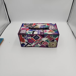 May include: A rectangular tissue box cover with a geometric pattern in various colors, including purple, green, pink, and white. The cover has a slot on top for tissue dispensing. The design features shapes like squares, triangles, and circles.