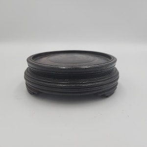 May include: A dark brown, round wooden display stand with a tiered base. The stand has a circular top and a series of carved, horizontal ridges on the base. The stand is designed to elevate and showcase decorative items.