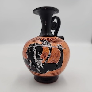 May include: A small, round terracotta vase with a black handle and spout. The vase features a black and white illustration of a figure with a spear. The vase is approximately 6 inches tall.