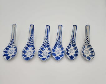 May include: A set of six ceramic soup spoons arranged in a row, each featuring a white base with a hand-painted blue floral and leaf pattern that extends from the bowl up the handle, showcasing a consistent design across all pieces.