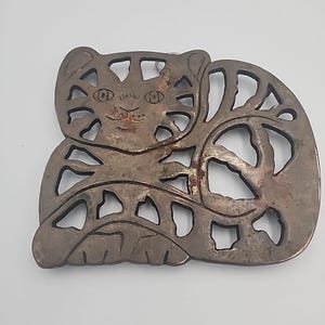 May include: A silver-coloured metal cat-shaped object with cut-out details. The cat has a smiling face and a striped pattern on its body. It is likely a decorative item or a piece of art.