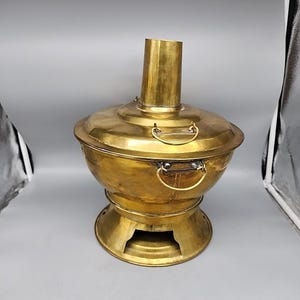 May include: A vintage brass hot pot with a cylindrical chimney and a round base. The pot has a lid with two handles and two side handles. The brass has a warm, golden colour.
