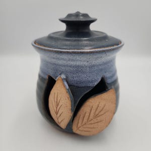 May include: A ceramic jar with a lid, featuring a dark blue and light blue speckled glaze. The body of the jar has two cut-out sections, each decorated with a raised, unglazed tan clay leaf applique that displays etched vein details.