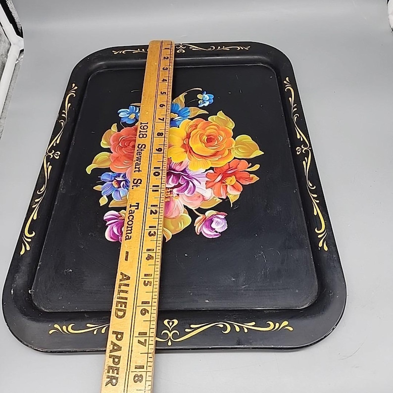 May include: Black rectangular serving tray with a floral design. The tray features a bouquet of colorful flowers in the center and gold trim along the edges. A wooden ruler is placed on top of the tray, measuring approximately 18 inches.