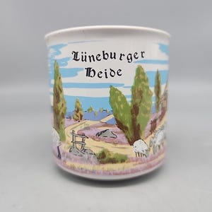 May include: White ceramic mug with a colorful landscape scene. The mug features a painted illustration of a rural landscape with trees, a road, and sheep. The text "Lüneburger Heide" is printed on the mug.