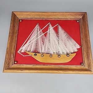 May include: A framed string art sailboat on a red background. The wooden boat is tan with gold-colored accents. White string forms the sails and rigging. The frame is made of light brown wood and is secured with small brass tacks.