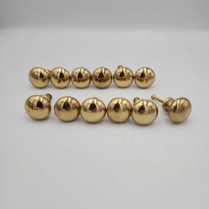May include: A set of twelve round, polished gold-tone metal cabinet knobs arranged in two rows. Each knob features a smooth, spherical head with a reflective surface and a short, threaded metal post extending from the back for mounting purposes.