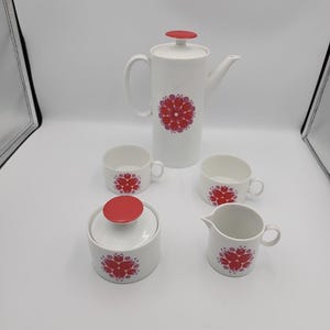 Puede incluir: Juego de café de cerámica blanca con diseño floral rojo. El juego incluye una cafetera alta, dos tazas, un azucarero con tapa y una jarra de crema. Las tapas y los detalles florales son rojos.