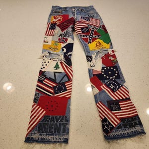 Può includere: Jeans in denim azzurro con finitura consumata e orli sfrangiati. I pantaloni sono riccamente decorati con varie toppe colorate, tra cui diversi disegni di bandiere americane storiche, simboli e la scritta TRAVEL AGENT.