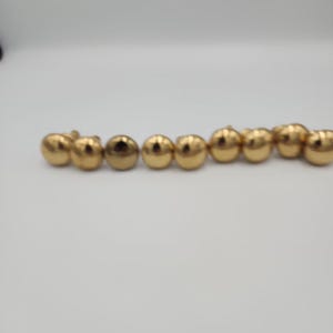 Puede incluir: Una colección de nueve botones metálicos pequeños de color dorado dispuestos en una fila horizontal. Cada botón presenta una superficie abovedada, lisa y pulida con un acabado reflectante, diseñada para su fijación en prendas o textiles.